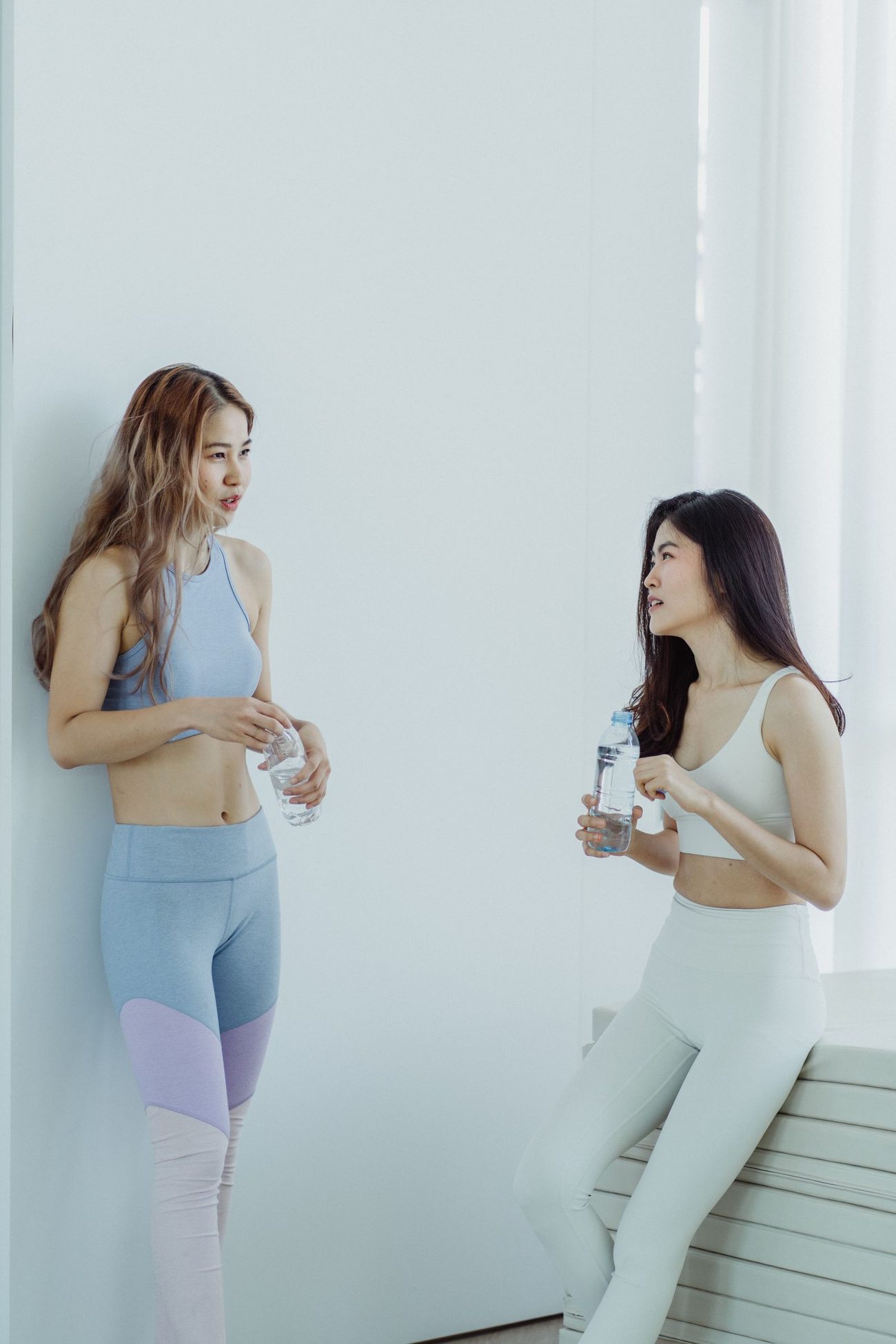 两位亚洲年轻女生穿着运动服，下课后靠着白墙聊天