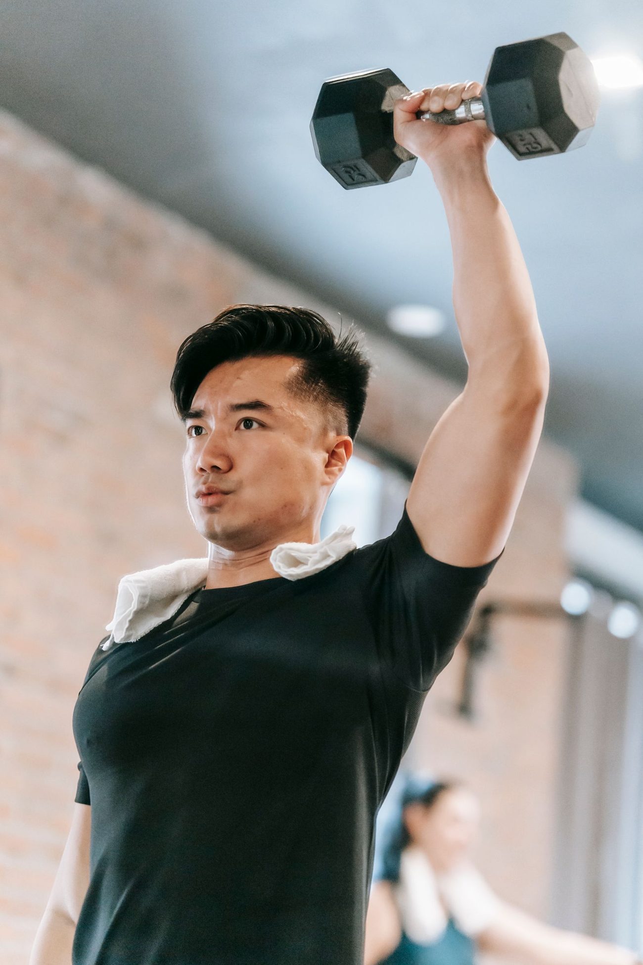 一位亚洲年轻男子在红砖墙健身房里专注地做哑铃推举