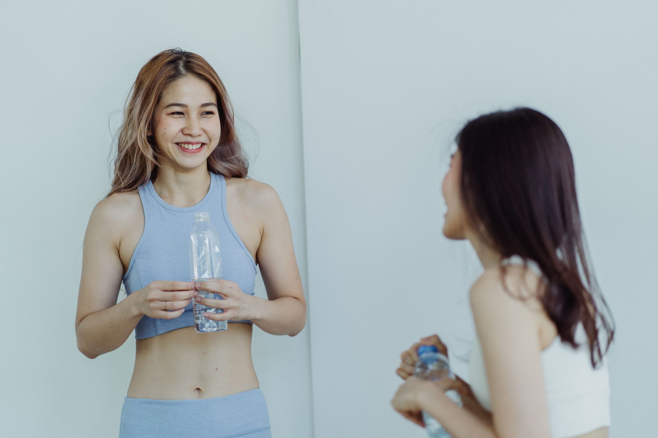 两位亚洲年轻女生穿着运动服，训练后拿着水瓶一起开心地笑